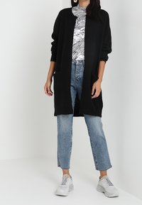 Czarny sweter o luźnym kroju z otwartym przodem, z dwiema przednimi kieszeniami. Nosi się go na wzorzystą bluzkę, w zestawieniu z jasnoniebieskimi dżinsami i białymi trampkami.