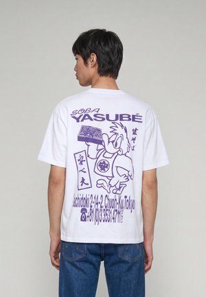 Ung mand med ryggen til iført en hvid T-shirt med lilla tegneserie-fugl, der holder soba-nudler og japansk tekst samt en Tokyo-adresse.