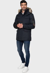 Marinblå parkasjacka med foder i huven, flera framfickor, ficka med dragkedja på ärmen, kombinerad med mörka jeans och vita sneakers.