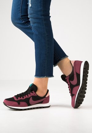 Person trägt bordeauxrote und schwarze Nike-Sneaker mit blauem Logo, kombiniert mit blauen, gekürzten Jeans mit fransigem Saum, steht auf weißem Boden.