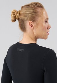 Černé dlouhé rukávy sportovního trička z hladké tkaniny. Má kulatý výstřih a malou značku na zadní straně. Přiléhavý střih.