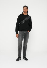 Zwarte langemouwen shirt, grijze spijkerbroek en zwarte sneakers met witte strepen. Het model draagt een zwarte schoudertas met een glanzende afwerking.