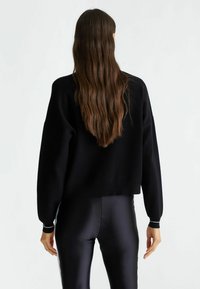 Maglione nero corto con maniche lunghe, polsini a costine e una texture liscia, abbinato a leggings neri lucidi.