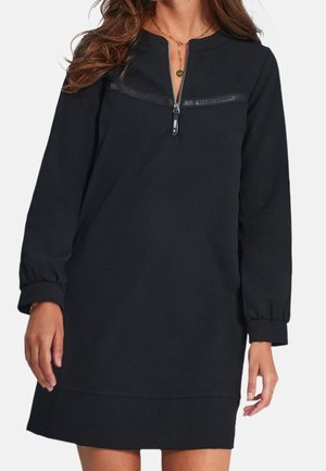 Femme portant une robe noire à manches longues avec une fermeture éclair à l'avant et un détail discret en texte sur la poitrine, associée à des colliers et un bracelet dorés.