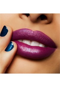 Primo piano di labbra truccate con un rossetto viola intenso e finitura lucida, accompagnato da uno smalto turchese su un dito vicino.
