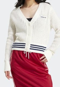 Bílý pletený cardigan s výstřihem do V, zapínáním na knoflíky a námořnicky modrým proužkovaným lemem. Nošený přes černý top a doplněný červenou sportovní sukní.