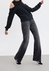 Personne portant un sweat-shirt noir à épaules dénudées, un jean noir délavé et des baskets noires et blanches, debout les bras écartés sur un sol gris.