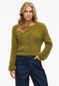 Frau mit blonden Haaren, die eine olivgrüne, flauschige Strickjacke und dunkelblaue Jeans trägt, steht mit einer Hand in der Tasche vor einem weißen Hintergrund.