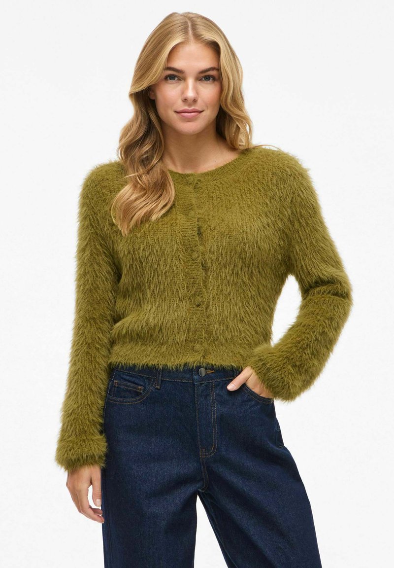 Frau mit blonden Haaren, die eine olivgrüne, flauschige Strickjacke und dunkelblaue Jeans trägt, steht mit einer Hand in der Tasche vor einem weißen Hintergrund.