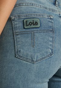 Vaqueros de mezclilla azul claro con un bolsillo trasero que muestra un parche rectangular verde con el texto "Lois" en negro. Detalle de costura estándar visible.