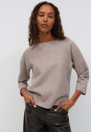 Donna con capelli ricci che indossa una maglia marrone chiaro a maniche lunghe e pantaloni in pelle neri con le mani nelle tasche, in piedi davanti a uno sfondo semplice.