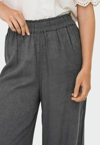 Šedé lněné kalhoty s elastickým pasem, zdobené sklady a volným střihem. Tkanina vypadá měkce s jemnou texturou.