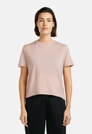 Kvinde med kort brunt hår iført en lyserød T-shirt med korte ærmer og sorte bukser, stående mod en hvid baggrund.