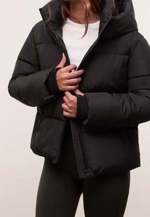 Persoon draagt een zwarte donzen jas met duimgaten, terwijl hij deze openritst over een wit shirt en donkere broek tegen een effen achtergrond.