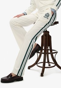 Hvide joggingbukser med grønne sidestrefe og et broderet logo. Sættet suppleret med mørk burgunder loafers på en træskammel.