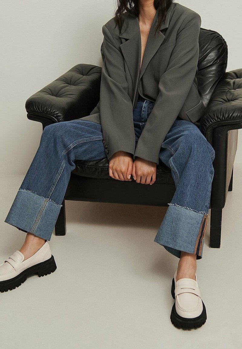 Personne assise sur un fauteuil en cuir noir, portant un blazer gris, un jean large retroussé et des mocassins blancs à semelles noires épaisses.