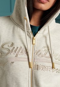 Hoodie beige chiaro con testo ricamato "SuperDry", cerniera dorata e dettagli in oro, dotato di cappuccio con coulisse e texture morbida.