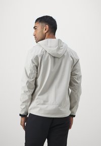 Giacca con cappuccio di colore grigio chiaro realizzata in un materiale liscio e resistente all'acqua; presenta polsini elasticizzati e un design leggermente aderente.