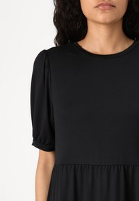 Vestuário preto com mangas curtas e bufantes com punhos enrolados, decote redondo e saia franzida. Feito de um tecido suave e elástico.
