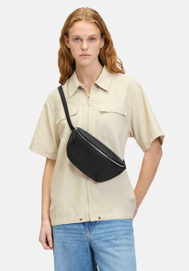 Schwarze Stoff-Crossbody-Tasche mit geschwungener Form, ausgestattet mit einem Reißverschluss und einem verstellbaren Trageriemen. Getragen über einem hellbeigen Hemd.