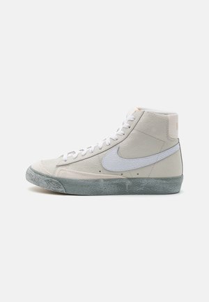 Höga sneakers i off-white med texturerad grå sula, vita snören och en vit Nike-swoosh på sidan, visad från utsidan.