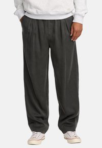 Dunkelgraue, plissierte Hosen mit lockerer Passform, aus weichem Stoff, mit Seitentaschen und kombiniert mit hellen Sneakers.