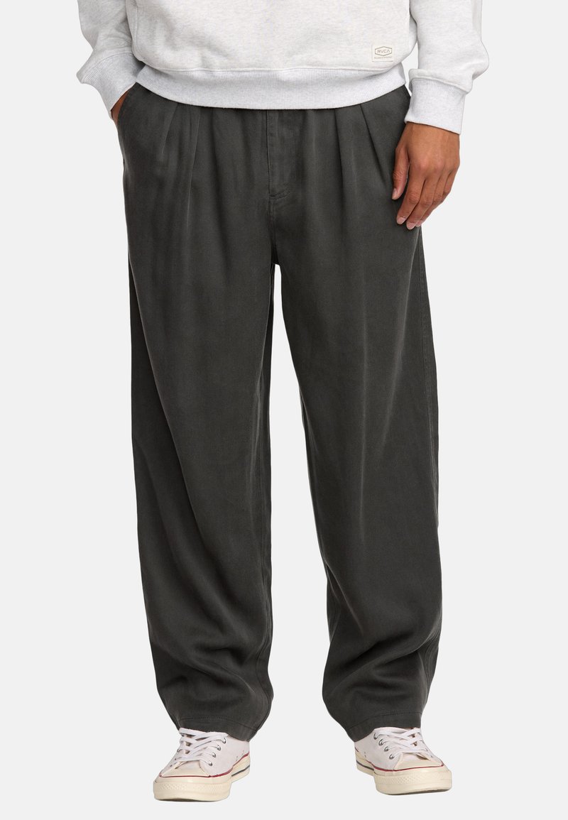 Dunkelgraue, plissierte Hosen mit lockerer Passform, aus weichem Stoff, mit Seitentaschen und kombiniert mit hellen Sneakers.