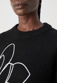 Schwarzer Strickpullover mit einem gerippten Rundhalsausschnitt. Auf der Vorderseite befindet sich eine weiße Blumenstickerei, die einen strukturierten Kontrast bietet.