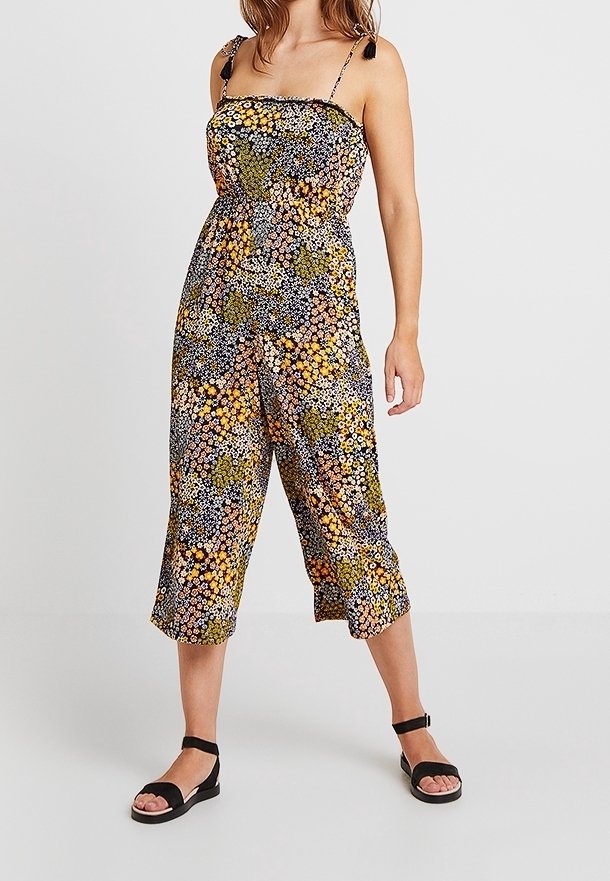 Femme portant une combinaison sans manches à fleurs avec des jambes larges raccourcies et des sandales noires à brides à la cheville, debout devant un fond uni.