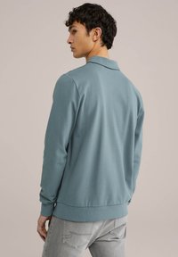 Svetlo modra polo majica z dolgimi rokavi in ovratnikom, ki ima gladko teksturo in rebraste manšete. Zadnja stran prikazuje minimalističen dizajn.