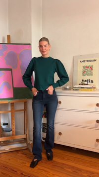 Top de algodão verde de mangas compridas com mangas baloon, combinado com jeans flare azul escuro e sapatos pretos de bico quadrado, de pé contra um fundo artístico.