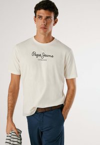 T-shirt en coton crème avec un col rond, arborant un logo noir imprimé "Pepe Jeans LONDON" sur la poitrine, associé à un pantalon bleu marine.