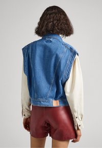 Giacca senza maniche in denim con una forma strutturata, di colore blu e dettaglio sul retro. Indossata sopra una camicia bianca a maniche lunghe e pantaloni corti rossi.