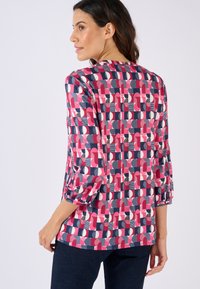 Blusa colorida con patrones geométricos en rosa, blanco, azul marino y negro. Mangas tres cuartos con acentos fruncidos, textura suave.