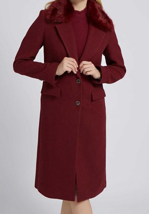 Kobieta w długim bordowym płaszczu z kołnierzem z futra, zapinanym na guziki w środku, pokazana od ramion do kolan na jednolitym tle.