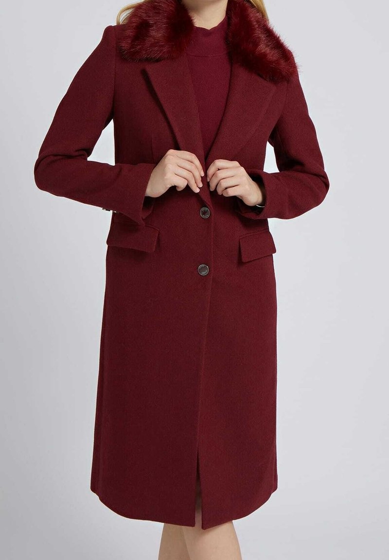 Femme portant un long manteau bordeaux avec un col en fourrure, boutonné au milieu, vue des épaules aux genoux sur un fond uni.