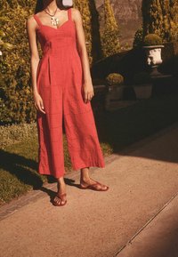 Tuta rossa in cotone con spalline sottili, scollatura quadrata e pantaloni fino al ginocchio. Texture con motivi ritagliati. Abbinata a ciabatte rosse.