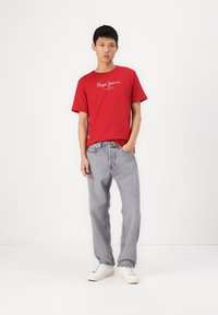 Camiseta de algodón roja con el logotipo "Pepe Jeans London", combinada con jeans de denim gris claro y zapatillas blancas. Diseño sencillo, ajuste relajado.