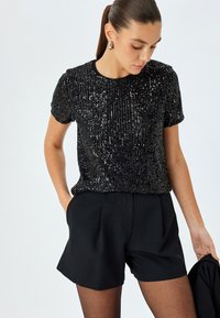 Zwarte korte mouw top van paillettenstof, met verticale strepen van pailletten. Gecombineerd met zwarte high-waisted shorts.