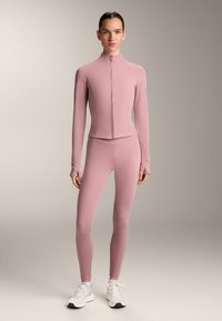 Tight rosa träningsset med jacka med hög krage och matchande leggings. Tillverkad av ett mjukt, stretchigt material. Vita sneakers.