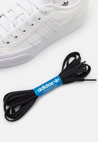 Bílé plátěné tenisky se třemi bílými pruhy, gumovou podrážkou a černými kulatými tkaničkami s modrým logem Adidas.