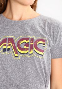 Grå bomulds T-shirt med et stort, slidtegn grafisk design med ordet "MAGIC" i gul, rød og sort, rund halsudskæring og korte ærmer.