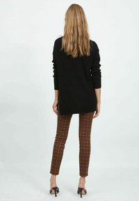 Schwarzer gerippter Pullover mit langen Ärmeln, kombiniert mit braunen karierten Hosen und schwarzen High-Heels, von hinten betrachtet.