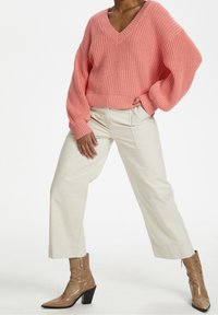 Maglione rosa lavorato a maglia spessa con scollo a V profondo, abbinato a pantaloni crema a gamba larga e stivali beige con tacco e punta affilata.