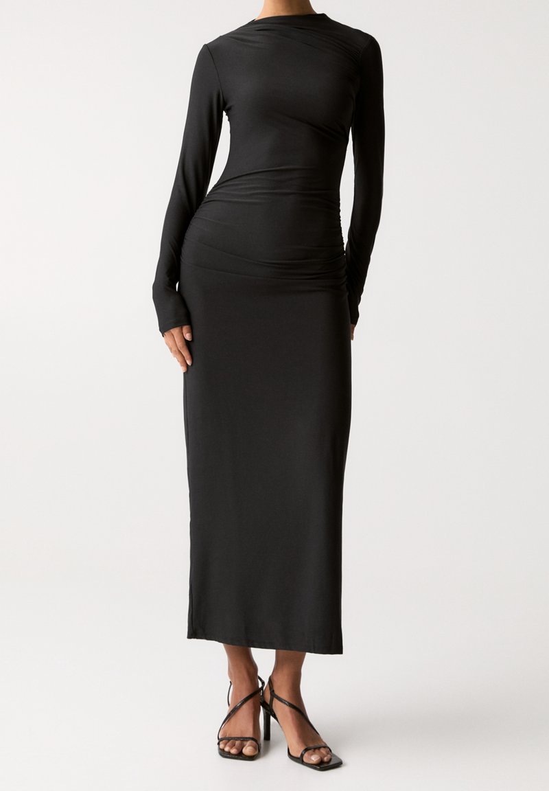 Femme portant une robe maxi noire cintrée à manches longues avec des trous pour les pouces et des sandales à talons hauts noires à brides sur fond blanc.