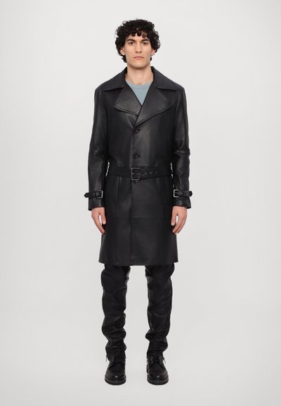 Manteau long en cuir noir avec un col prononcé, boutons croisés, taille ceinturée et poignets détaillés, associé à un pantalon noir ajusté.