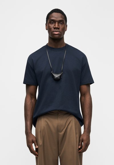 Tmavě modré bavlněné tričko s krátkými rukávy a kulatým výstřihem, kombinované s khaki kalhotami a malým černým náhrdelníkem v podobě pouzdra.