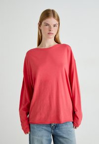Jongere met lang steil haar, draagt grote ringoorbellen, een losse koraalkleurige blouse met lange mouwen en lichtblauwe jeans, staande tegen een effen achtergrond.