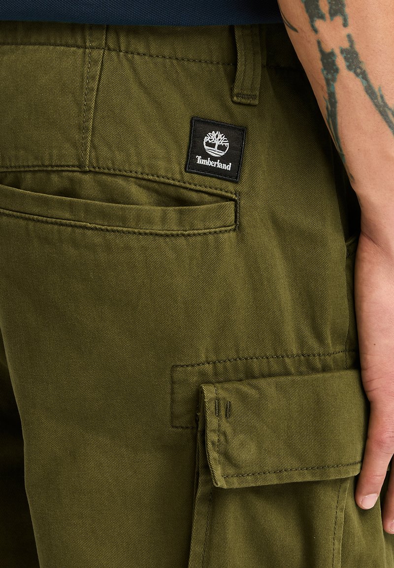 Oliwkowe spodnie cargo z wieloma kieszeniami, czarną naszywką z logo Timberland oraz widocznymi detalami ściegów z wytrzymałego materiału.