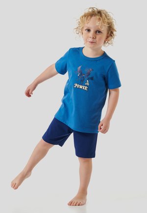 Blaues Baumwoll-T-Shirt mit kurzen Ärmeln und einem grafischen Design, das einen Charakter zeigt, kombiniert mit marineblauen Shorts. Kind steht auf einem Bein.
